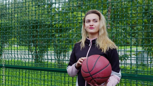 Attractive blonde woman in tracksuit holding ball, going to play basketball or volleyball. Fit girl stands on fenced street sports ground. Lady decided to play sport. Female basketballer, hoopster photo