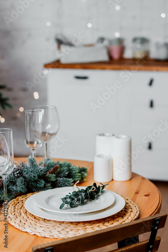 Round table decorated for Christmas and New Year. Christmas tree twigs on the table. Plates and wine glasses on the table. Kitchen set and flowers on the background. Christmas content.