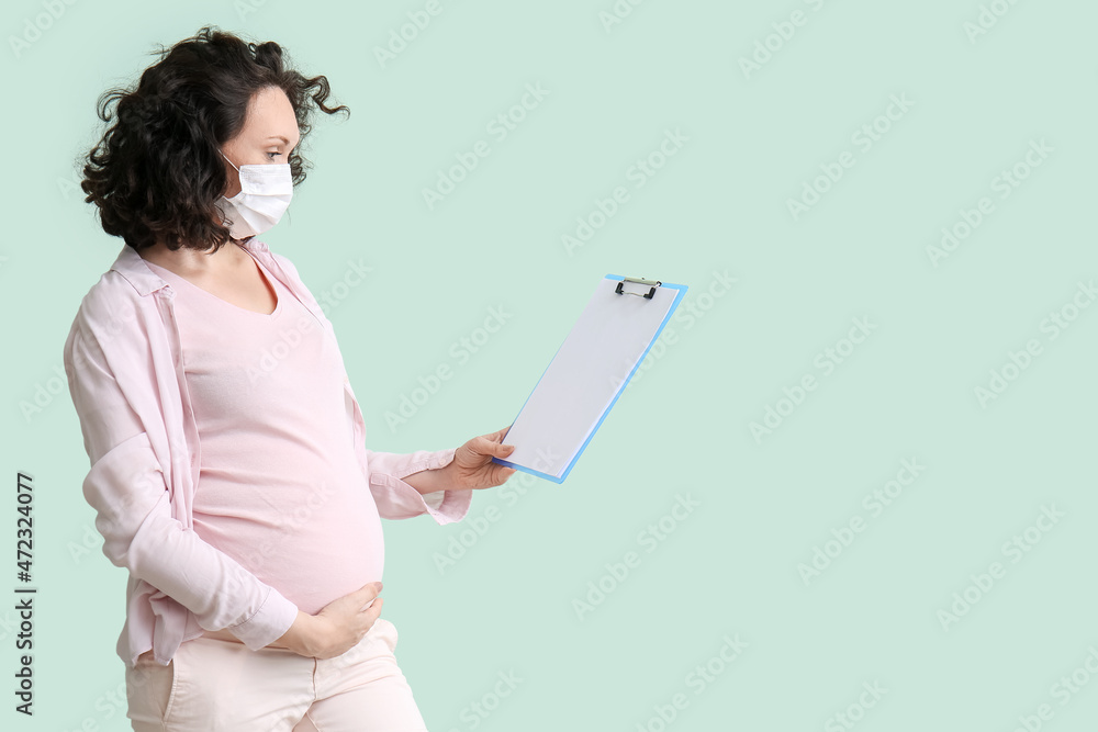 Young pregnant woman in medical mask with clipboard on color background Young pregnant woman in medical mask with clipboard on color background