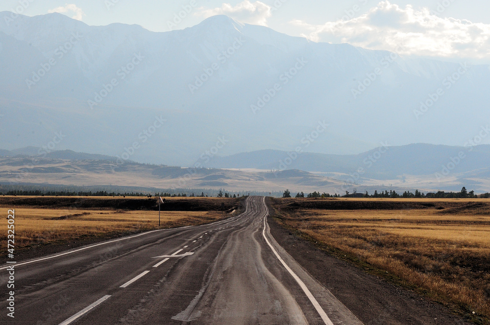 A winding road going through the autumn steppe to meet hills and snow-covered mountain peaks.