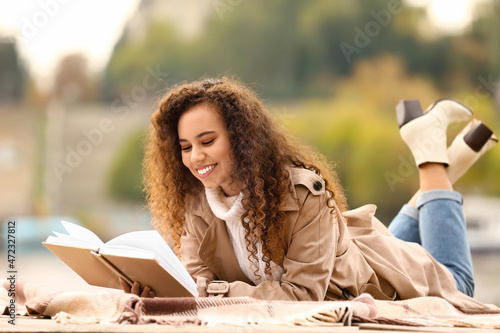 Beautiful African-American woman reading interesting book outdoors