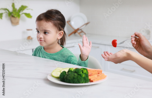 Mother feeding her daughter in kitchen, closeup. Little girl refusing to eat vegetables photo