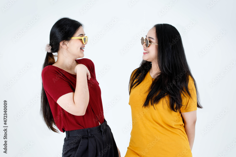 Studio shot of two young Asian cool fashionable stylish female friends in casual tshirt outfit wearing fashion vintage sunglasses standing smiling looking at each other eyes on white background Studio shot of two young Asian cool fashionable stylish female friends in casual tshirt outfit wearing fashion vintage sunglasses standing smiling looking at each other eyes on white background