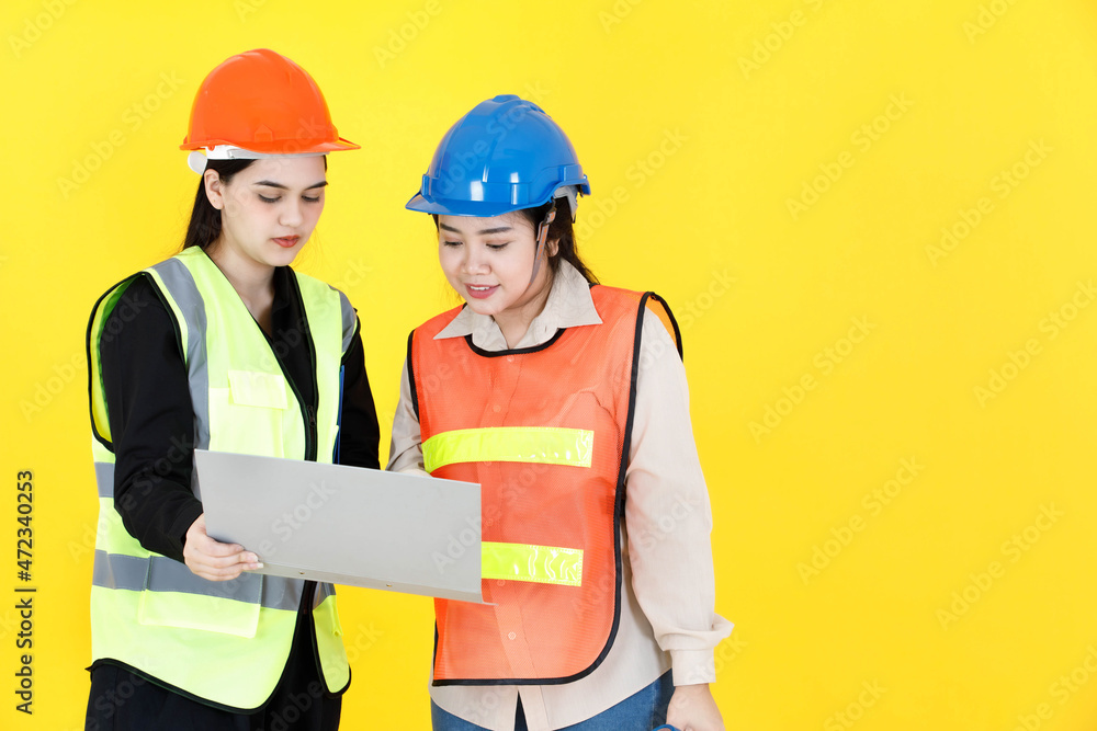 Studio shot of two Asian female professional engineering manager foreman wearing hard helmet and reflective safety vest holding paperwork document file folder discussing together on yellow background Studio shot of two Asian female professional engineering manager foreman wearing hard helmet and reflective safety vest holding paperwork document file folder discussing together on yellow background