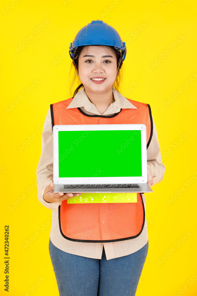 Portrait studio shot of Asian female professional engineering foreman manager in hard helmet and reflect safety vest hold blank green screen laptop computer for text copy space on yellow background Portrait studio shot of Asian female professional engineering foreman manager in hard helmet and reflect safety vest hold blank green screen laptop computer for text copy space on yellow background