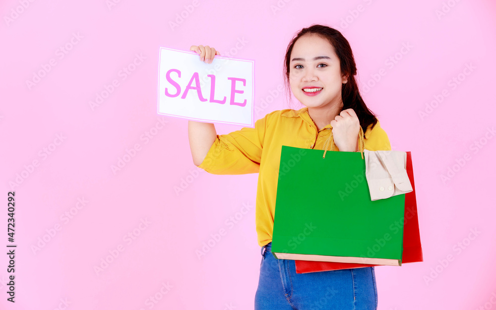 Fototapeta premium Portrait studio shot Asian young happy female shopper wearing casual outfit carrying colorful shopping bags holding showing sale discount banner paper sign look at camera smiling on pink background