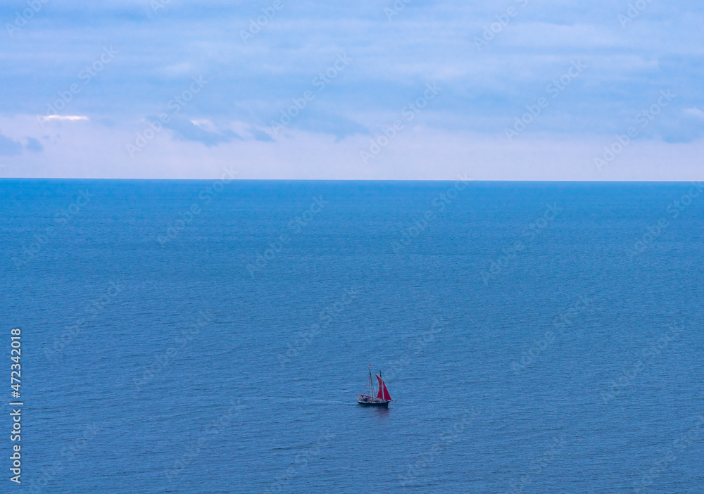 A lonely yacht with scarlet sail sails on the blue sea in the distance in cloudy weather