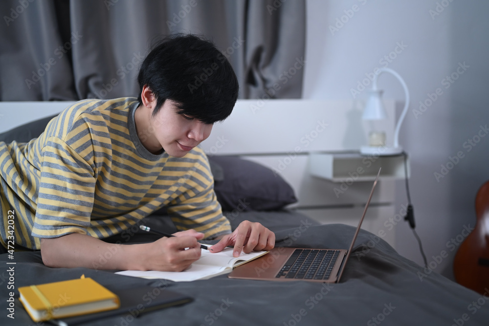 Smiling asian man lying on bed and studying online with laptop computer. Smiling asian man lying on bed and studying online with laptop computer.
