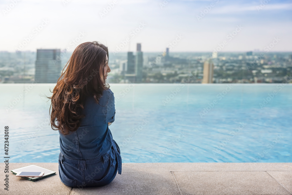 Luxury vacation woman relaxing in infinity swimming pool on summer travel. Asian girl tourist relax after working on Tablet, Smartphone looking back at camera Blurred background Skyscraper, Sky,Clouds