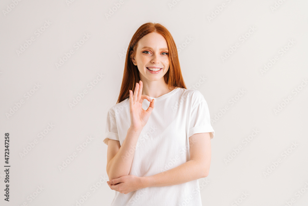 Portrait of smiling pretty redhead young woman showing okay gesture looking at camera, standing on white isolated background in studio. Fronv view of cheerful attractive female showing ok sign gesture Portrait of smiling pretty redhead young woman showing okay gesture looking at camera, standing on white isolated background in studio. Fronv view of cheerful attractive female showing ok sign gesture