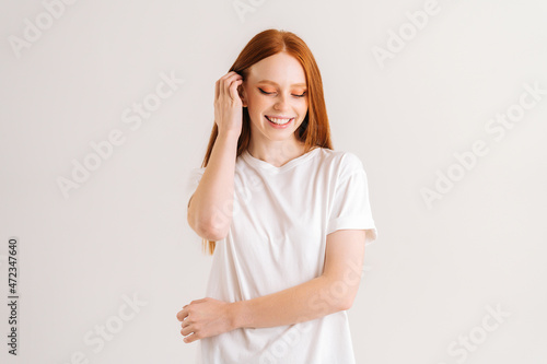 Portrait of charming smiling young woman touching playfully red hair looking down, on white isolated background in studio. Happy atractive redhead lady flirting with you.