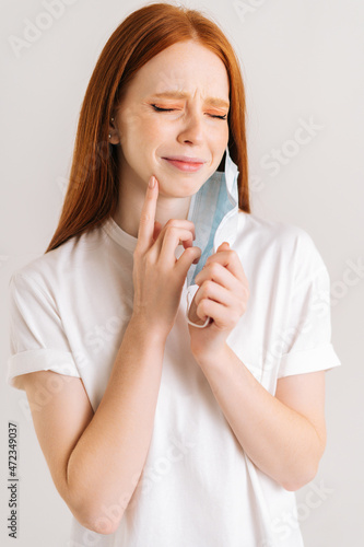 Close-up vertical shot of suffering young woman taking off disposable protective face mask, pointing finger to chin, touching swollen, spot, maskne, scar, skin allergy, on white isolated background.