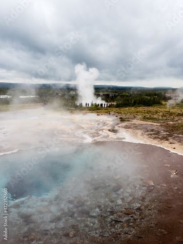 Steaming colorful hot spring pool with Strokkur geyser at the background in Geysir geothermal area, Iceland