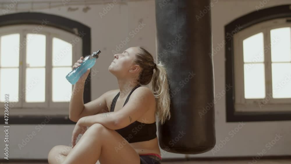 Side view of athletic woman drinking isotonic in gym. Medium shot of tired Caucasian sportswoman recovering after kickboxing training, sitting on floor and resting. Sport, relaxation concept Side view of athletic woman drinking isotonic in gym. Medium shot of tired Caucasian sportswoman recovering after kickboxing training, sitting on floor and resting. Sport, relaxation concept