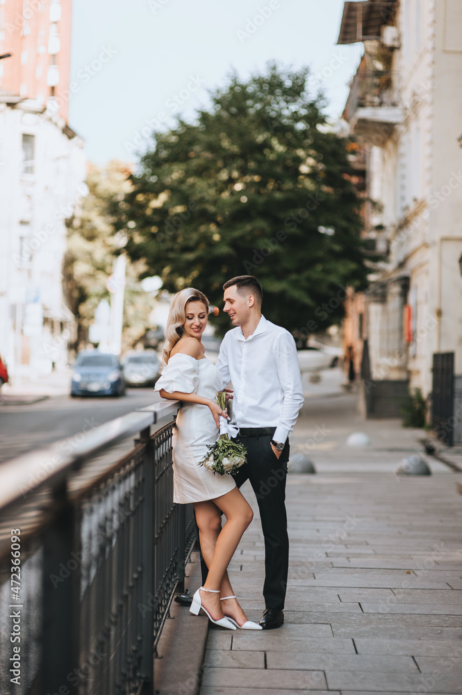 A young groom and a beautiful blonde bride in a white dress are hugging, standing on the alley in the city on the street. Wedding photography.