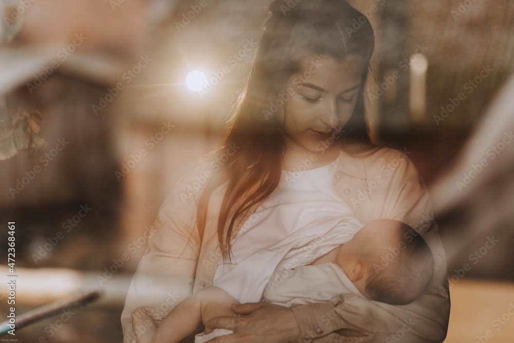 A young mother holds her baby near her breast at home in a dark bedroom by the window. The concept of breastfeeding, a photo of a real family, not a model A young mother holds her baby near her breast at home in a dark bedroom by the window. The concept of breastfeeding, a photo of a real family, not a model