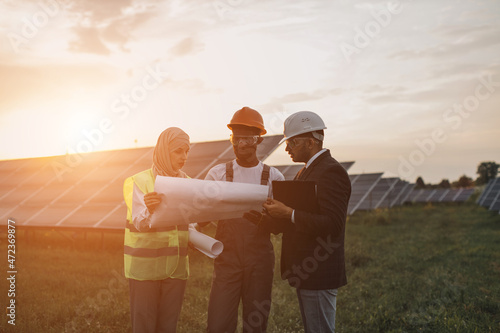 Group of three multiracial people in safety helmets having working meeting among solar station. Technician with engineers standing outdoors with blueprints in hands.