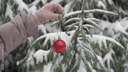 Female hands in winter gloves hang a red ball, a Christmas tree toy on a Christmas tree in a snowy winter forest. Fluffy snow is falling