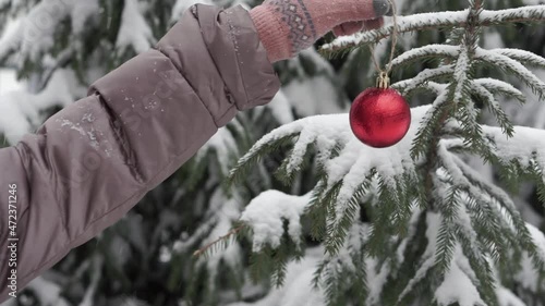 Female hands in winter gloves hang a red ball, a Christmas tree toy on a Christmas tree in a snowy winter forest. Fluffy snow is falling
