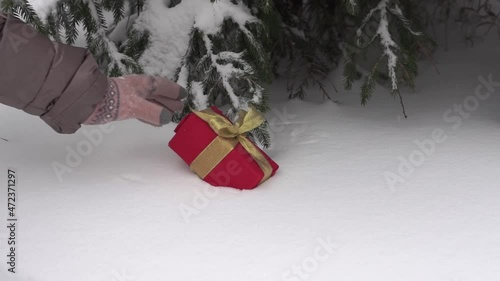 Female hands in winter gloves put a Christmas gift box under the tree in a snowy winter forest. Fluffy snow is falling.