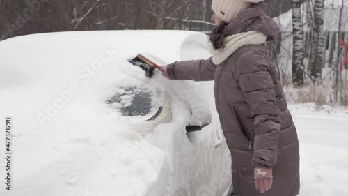 A middle-aged woman cleans car windows from snow in the winter season. Snow drifts