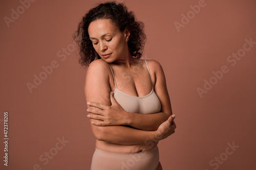 Confident serene beautiful curly woman with imperfect body, cellulite and stretch marks after childbirth, in beige underwear, looking down, hugging herself on a colored background with copy ad space