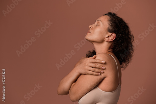 Side view of a beautiful middle aged Hispanic woman in beige underwear hugging herself, lifting her head, posing with closed eyes against colored background with copy ad space. Body positivity concept photo