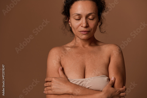 Close-up portrait of a serene Hispanic woman posing with closed eyes, hugging herself against beige background with copy ad space.