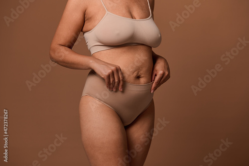 Close-up body of overweight woman with stretch marks and saggy belly after pregnancy, isolated on beige background with copy space for ads. Concept of love and acceptance, body positive.