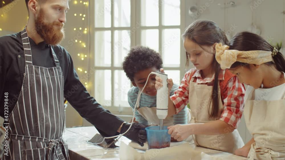 Little Caucasian girl using hand blender with help of male chef while cooking with diverse kids during culinary class Little Caucasian girl using hand blender with help of male chef while cooking with diverse kids during culinary class