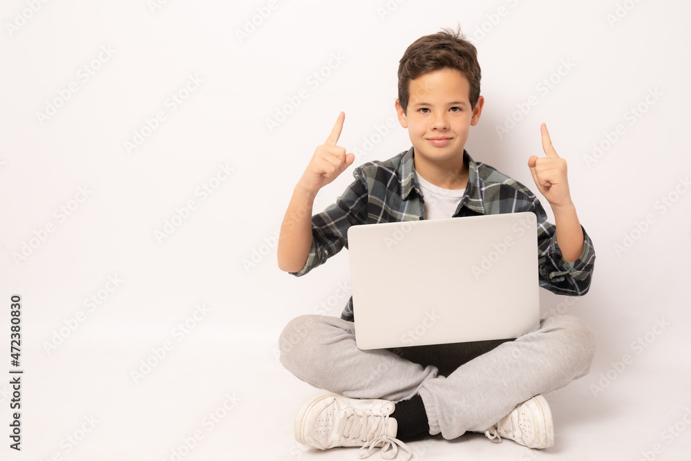 Smiling cute boy wearing plaid shirt sitting on floor holding laptop computer pointing fingers up isolated over white background. Smiling cute boy wearing plaid shirt sitting on floor holding laptop computer pointing fingers up isolated over white background.