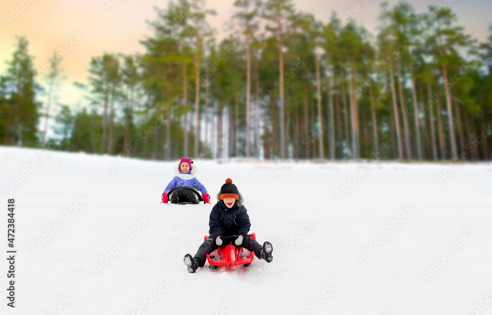 childhood, sledging and season concept - happy little children sliding on sleds down snow hill in winter over snowy forest or park background