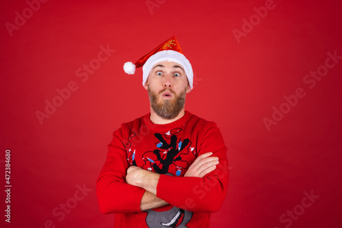 Red-bearded man in santa hat and christmas sweater on red background posing cheerfully, smiling excitedly