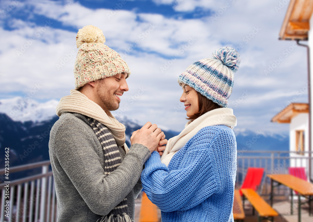 Fototapeta premium travel, tourism and winter holidays concept - happy romantic couple in knitted hats and scarves holding hands over mountains and ski resort background