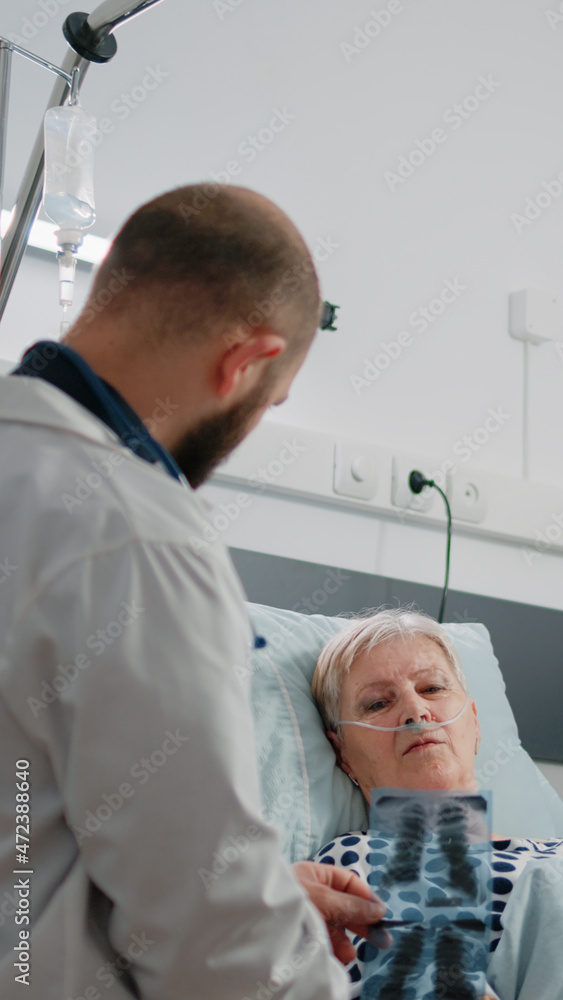 Doctor showing diagnosis of radiography to patient with illness laying in hospital ward bed. Medic holding x ray scan for retired woman while nurse giving assistance and support. Doctor showing diagnosis of radiography to patient with illness laying in hospital ward bed. Medic holding x ray scan for retired woman while nurse giving assistance and support.