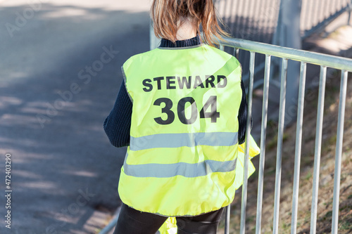 Young student girl participates as a volunteer in ensuring the security of a city event. Standing near the fence with the inscription Steward on the jacket photo