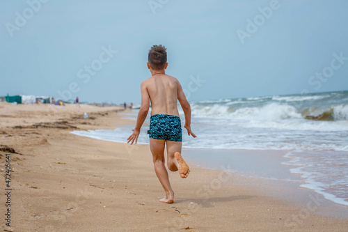 The boy runs to the sea on the beach. View from the back. Cloudy summer day
