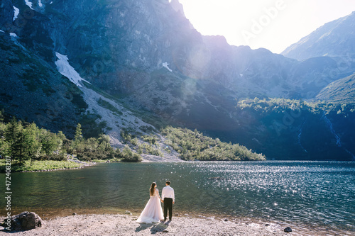 The moment of the wedding  the newlyweds look at the lake. Rear view of young amazing bride and groom looking at the lake in the mountains.