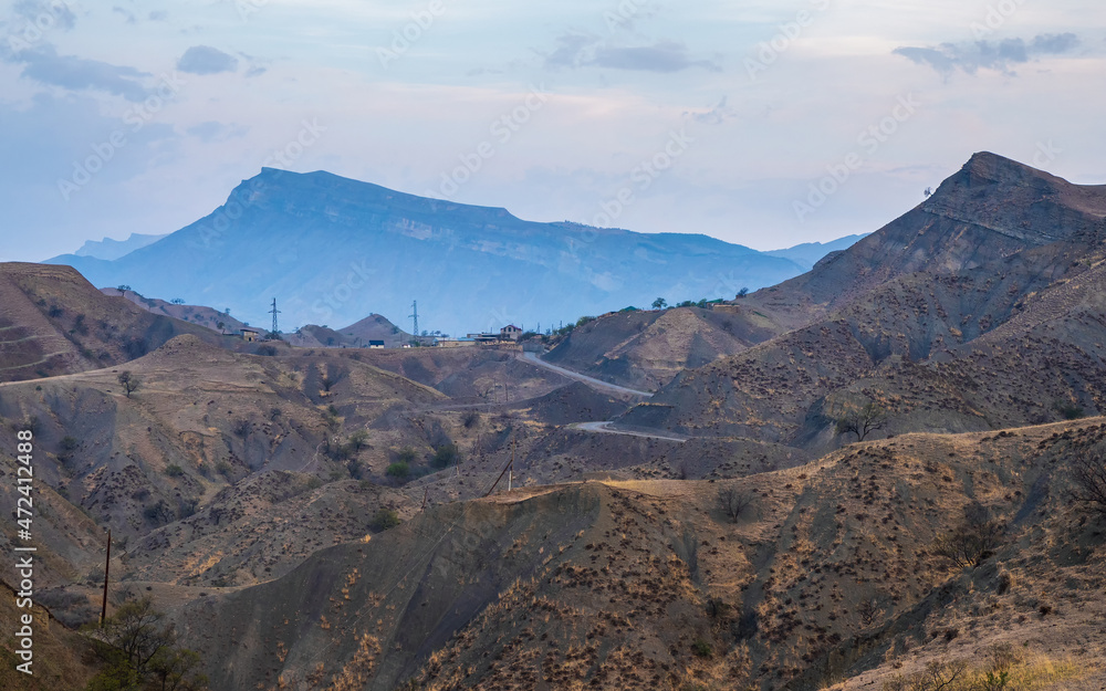 Panorama of the evening mountains with a complex landscape. Mountain serpentine in the dawn sunlight. Dangerous narrow dirt mountain road through the hills to a high-altitude village.