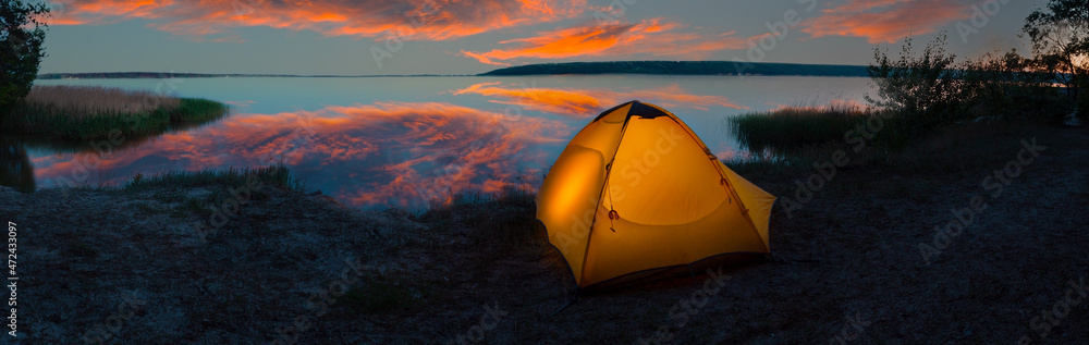 Orange tourist tent illuminated from inside on the lakeside under dramatic sky