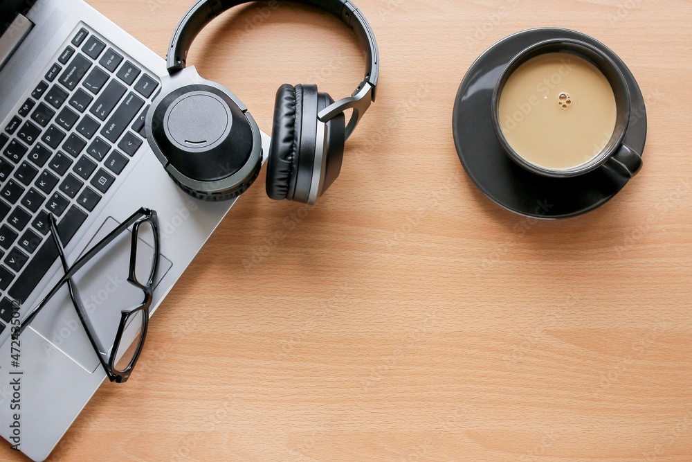 View from above of black headphone, and eyeglasses on the laptop, a cup of coffee on the wooden table. Work from home concept View from above of black headphone, and eyeglasses on the laptop, a cup of coffee on the wooden table. Work from home concept