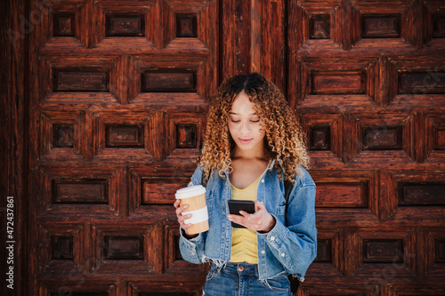 Curly haired woman holding reusable cup using smart phone by wooden door