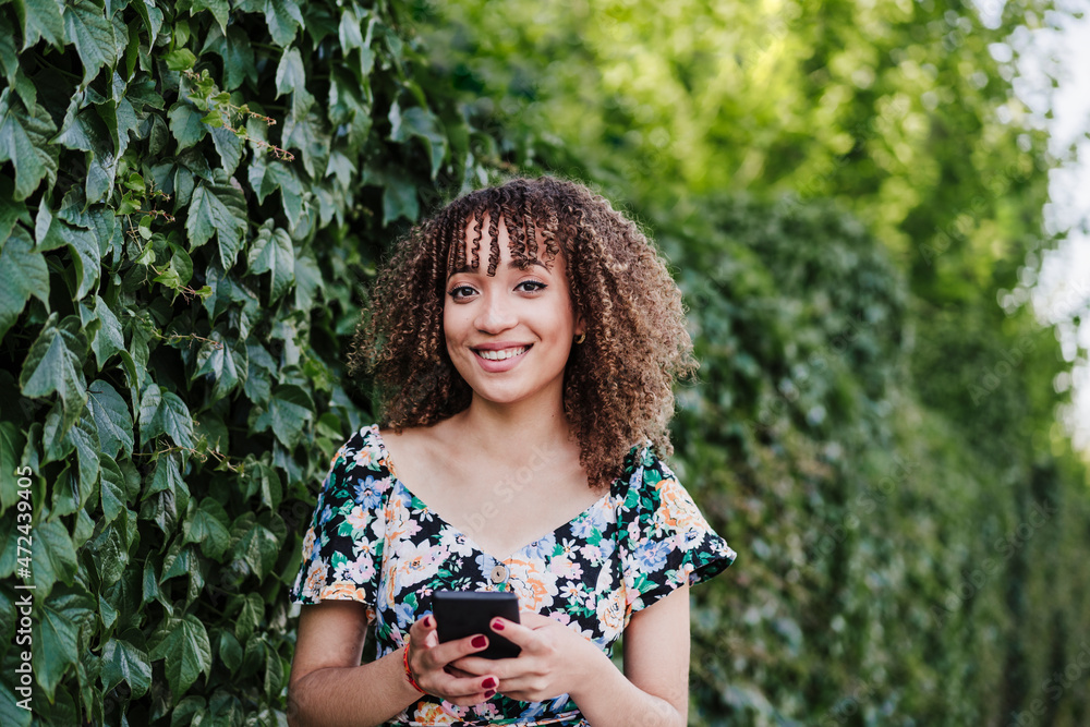 Smiling young woman holding smart phone while standing by ivy plant Smiling young woman holding smart phone while standing by ivy plant