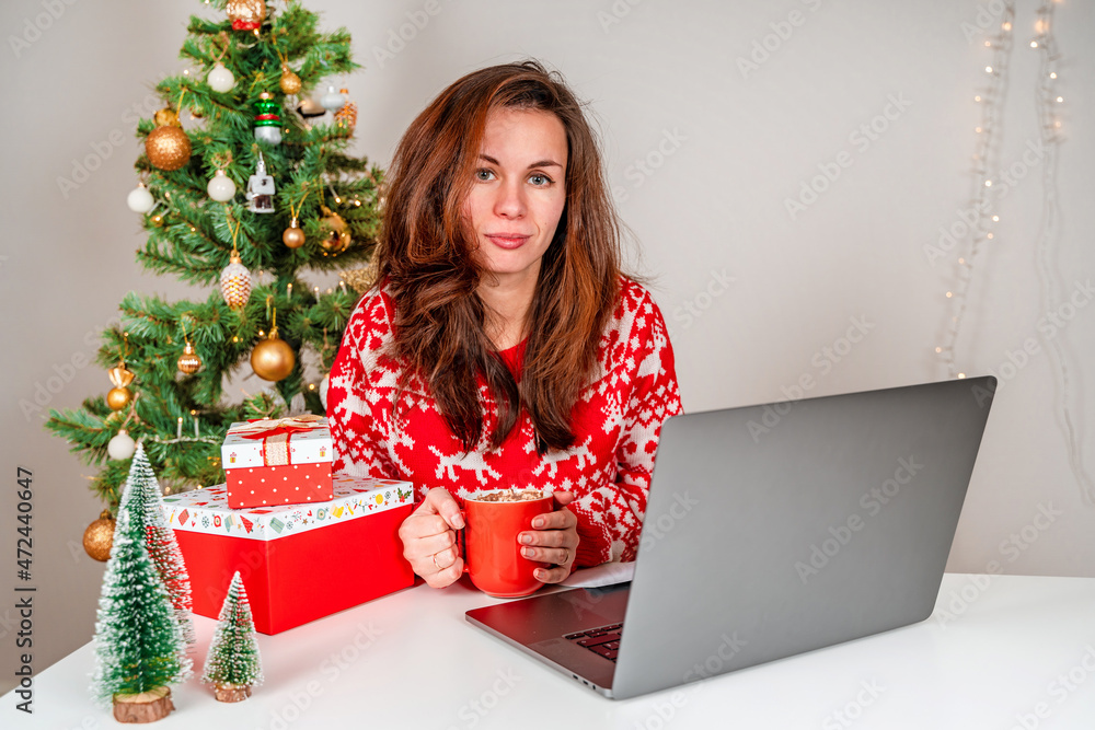 A young woman in a red sweater uses a laptop during the Christmas holidays and New Year. The concept of online shopping and communication A young woman in a red sweater uses a laptop during the Christmas holidays and New Year. The concept of online shopping and communication