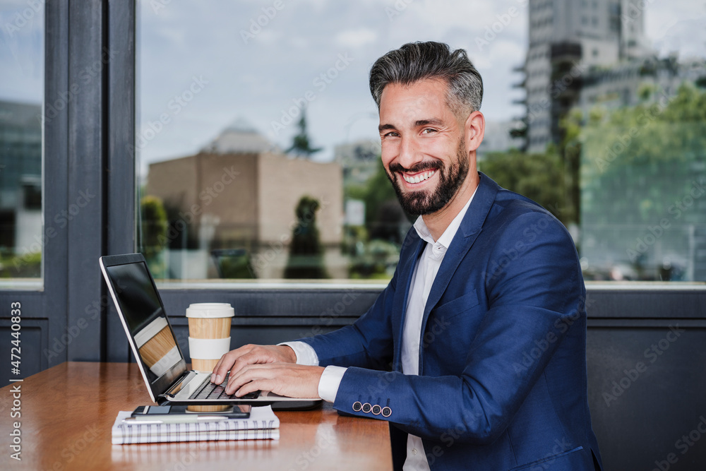 Smiling businessman with laptop sitting by window at cafeteria Smiling businessman with laptop sitting by window at cafeteria