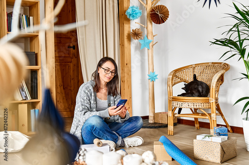 Woman using cell phone and play with black cat while wrapping christmas gifts in craft recycled paper in the room with alternative christmas tree and paper christmas decoration.Winter holidays photo