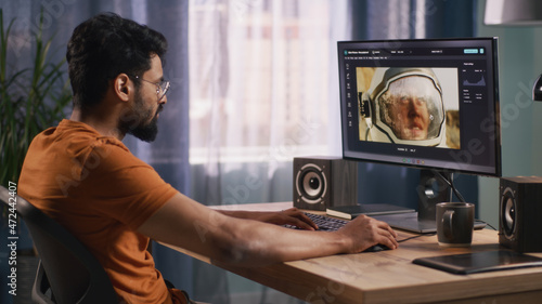 Side view of bearded Indian man using retouching software on computer to edit picture of cosmonaut during work in home office