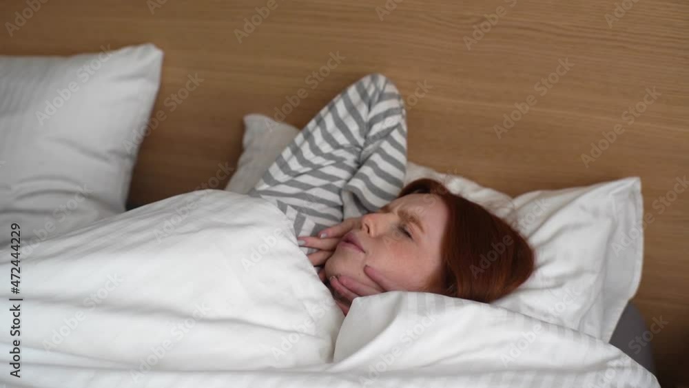 Cheerful young woman waking up, opening eyes while lying in comfortable white bed at home and looking to window. Happy beautiful redhead female stretching enjoying early good morning in cozy bedroom. Cheerful young woman waking up, opening eyes while lying in comfortable white bed at home and looking to window. Happy beautiful redhead female stretching enjoying early good morning in cozy bedroom.