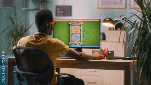 African American male gamer playing videogame on computer while sitting at table in evening at workplace
