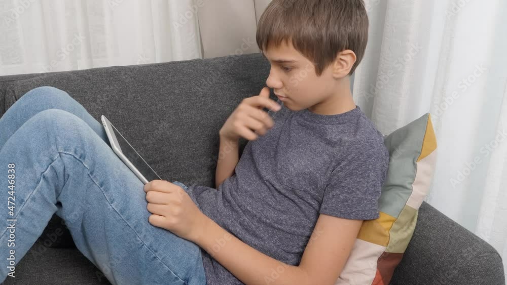 Teenage boy using digital tablet computer sitting on couch at home. Kid using application for learning, playing games, browsing internet, looking video, studying online Teenage boy using digital tablet computer sitting on couch at home. Kid using application for learning, playing games, browsing internet, looking video, studying online